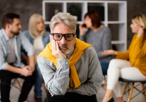 En eldre mann ser trist ut i en gruppeterapitime, omgitt av voksne som diskuterer psykiske helseproblemer. Viser psykiske helseproblemer hos voksne.