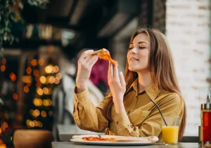 En kvinne koser seg med et pizzastykke på en restaurant – et bilde relatert til temaet spiseforstyrrelser og overspising, som illustrerer forholdet mellom mat og følelser.