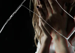 Kvinne skjuler ansiktet bak hendene med knust glass i forgrunnen, et sterkt visuelt uttrykk for følelsene knyttet til angst og traumer.