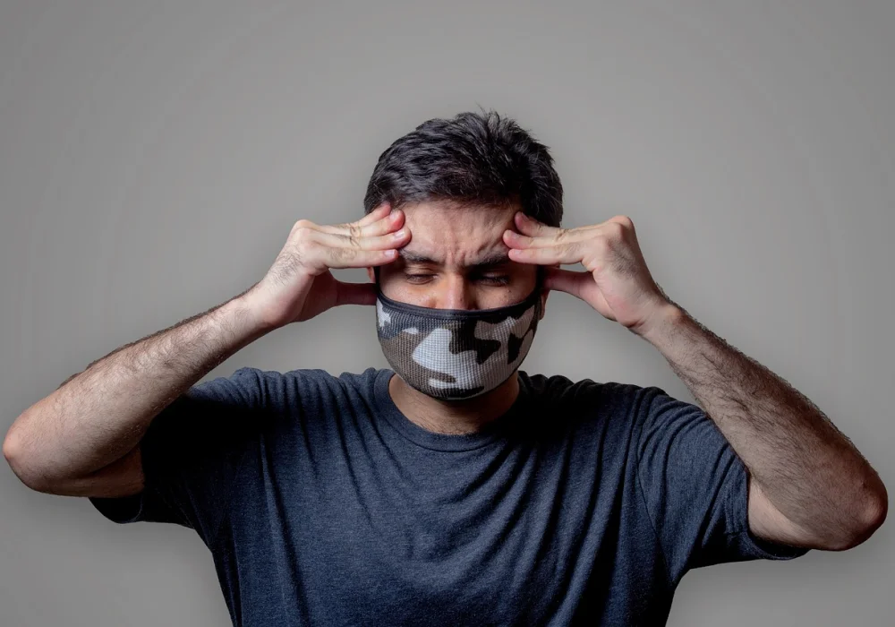 Mann med munnbind som holder hodet med begge hender og viser ubehag – illustrerer Spenningshodepine og stress i hverdagen.